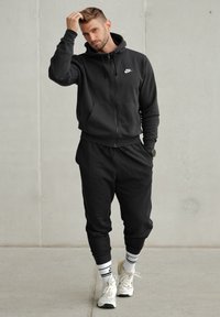Glugă Nike neagră cu fermoar, dotată cu buzunare tip kangaroo, asortată cu pantaloni jogger. Purtați cu adidași albi și șosete cu dungi. Design minimalist.