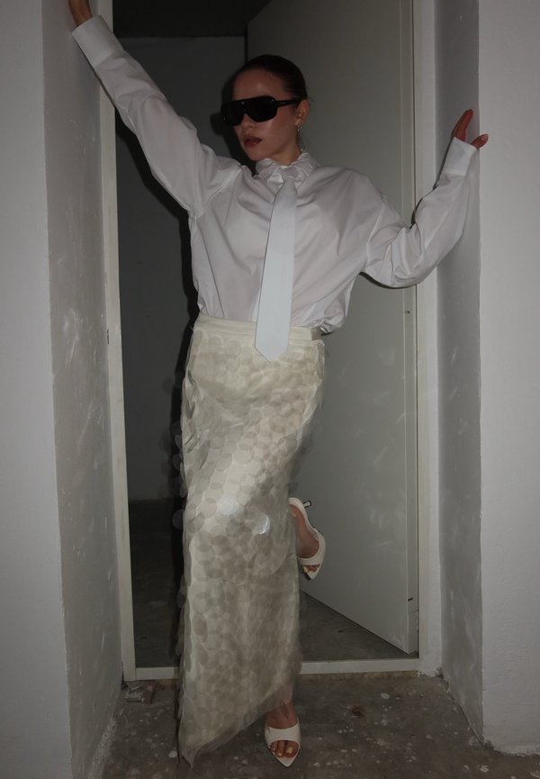 Persona que lleva una camisa blanca de gran tamaño con una corbata larga, falda blanca texturizada, tacones blancos y gafas de sol negras, posando en una puerta estrecha.