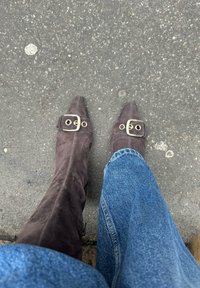 Rjave suede čevlje z ostrim nosom in velikim zlatim zaponko, v paru z modrimi kavbojkami, ki počivajo na temni asfaltni površini.
