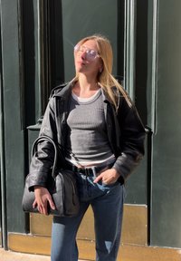 Crna kožna jakna preko sive rebraste majice, plave hlače visokog struka i crna torbica. Svijetle sunčane naočale odražavaju sunčevu svjetlost.