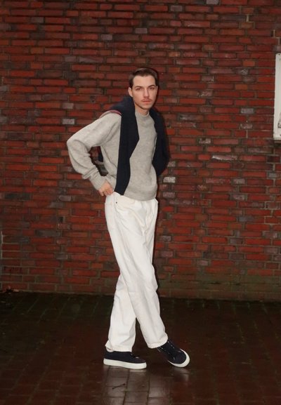 Suéter gris, chaleco marino colgado sobre los hombros, pantalones blancos y zapatos oscuros. De pie contra una pared de ladrillo rojo texturizada.