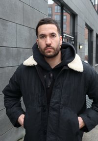 Homme aux cheveux courts et foncés avec une barbe, portant un manteau noir à col en fourrure blanche, debout à l'extérieur contre un mur carrelé gris dans un cadre urbain.