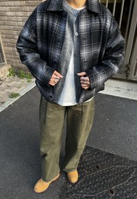 Personne portant une veste à carreaux noire et blanche sur un cardigan gris et une chemise blanche, un pantalon vert olive, des bottes jaunes et plusieurs bagues en argent.