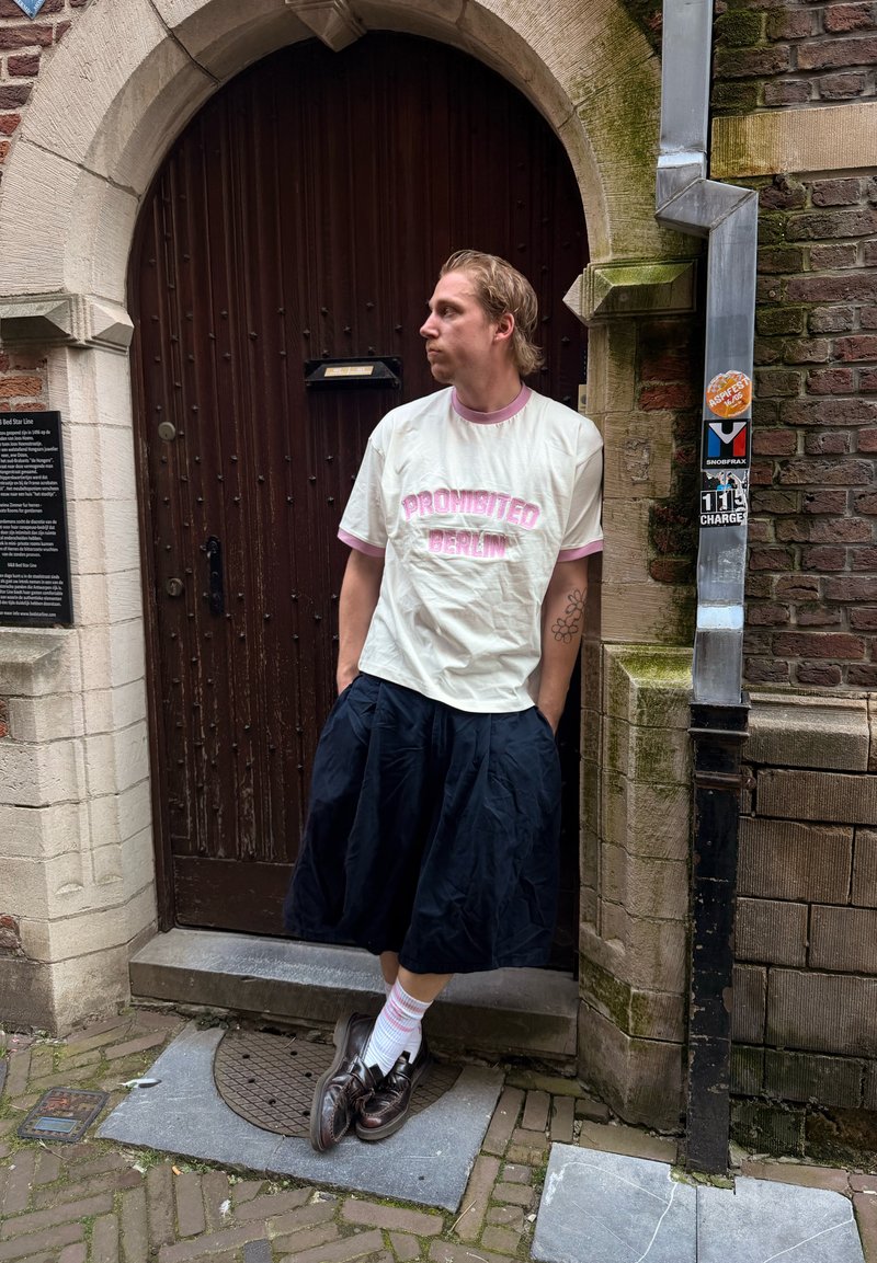 Jeune homme avec tatouage, portant une chemise blanche "Prohibited Berlin" et une jupe noire, appuyé contre une porte en bois arquée dans une ruelle en briques.