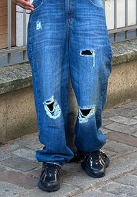 Jeans en denim délavé de bleu moyen avec deux grandes déchirures à l'avant. Coupe ample avec poches, assortis à des baskets noires.