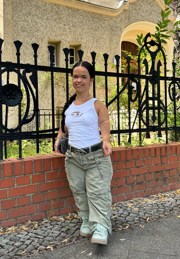 Bílý top bez rukávů s výřezem, olivové cargo kalhoty s kapsami a světle zelené platformové tenisky; stojí u cihlové zdi a plotu.