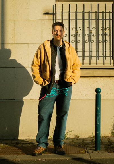 Chaqueta amarilla con capucha sobre una camisa negra con estampado, vaqueros azules con diseño turquesa, accesorio de cuentas rojas y zapatos de estampado de leopardo. De pie junto a una pared.