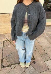 Person som bär en grå knappad cardigan, en tie-dye t-shirt, rivna ljusblå jeans, gröna slip-on skor och två halsband, stående på ett kaklat golv.