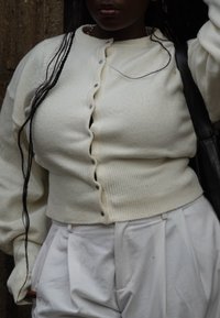 Gilet blanc court avec une texture côtelée et des détails de boutons ronds, associé à un pantalon blanc ample. Une sangle de sac noire est visible.