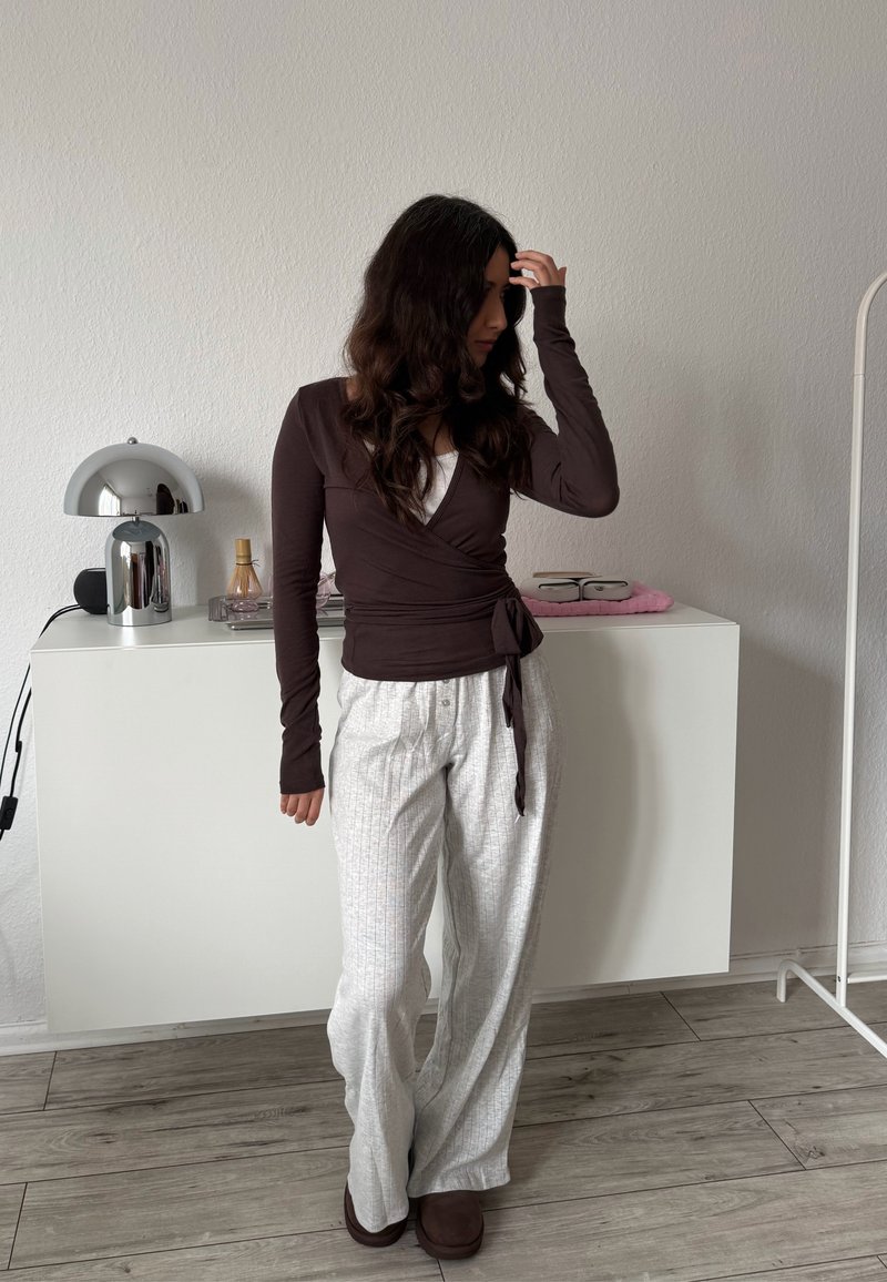 Žena s dlhými tmavými vlasmi si upravuje vlasy, má na sebe tmavo hnedý zavinovací top a široké biele nohavice s prúžkami, stojí v minimalistickej miestnosti pri bielom skrini.