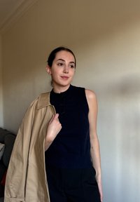 Námořnický bezrukávový top s vysokým límcem a detaily knoflíků, doplněný béžovým oversized sakem. Pozadí tvoří světlá zeď a pohovka.