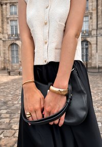 Black leather handbag with curved shape, worn with a textured cream top featuring gold buttons. Accessories include a gold bangle and a ring.