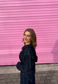 Jeans jakna temno modre barve, z ovratnikom in bakerasto opremo, postavljena ob svetlo roza steno.