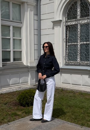 Mujer que lleva una camiseta negra, pantalones blancos, gafas de sol y zapatos negros, sosteniendo un bolso negro mientras está de pie afuera cerca de un edificio blanco con ventanas.