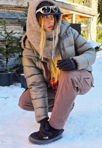 Donkergroen olijfgroene puffy jas, bruine cargobroek, grijze gebreide sjaal, zwarte handschoenen en zwarte North Face laarzen, tegen een besneeuwde achtergrond.