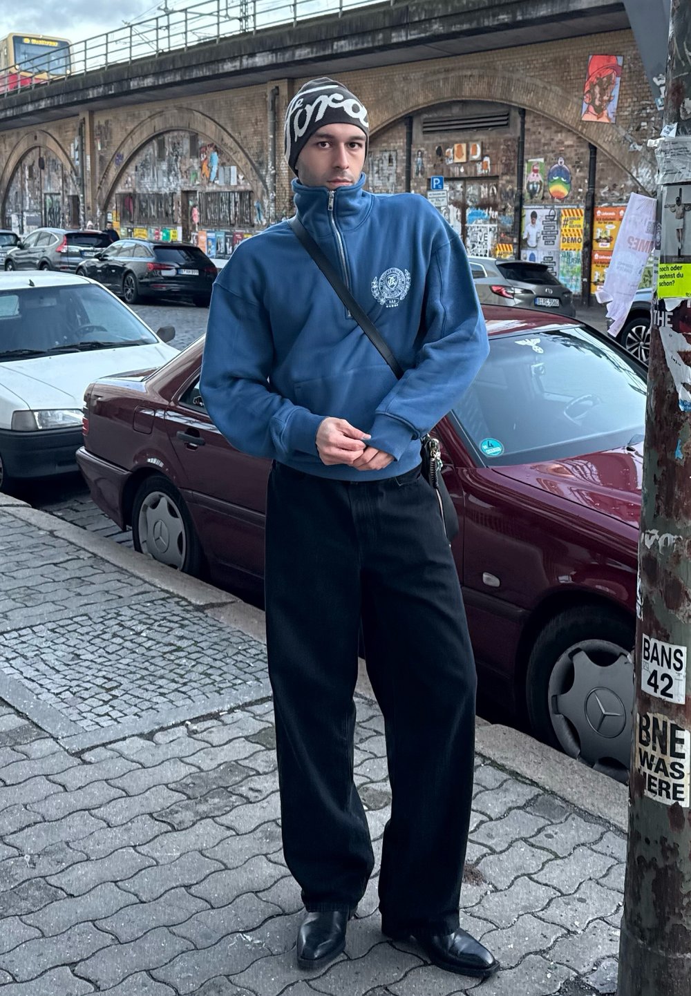 Μπλε φούτερ με φερμουάρ και λογότυπο, μαύροι πλατείς παντελόνες και μαύρα μυτερά παπούτσια. Αστική τοποθεσία με σταθμευμένα αυτοκίνητα.