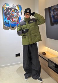 Νέος άνθρωπος με πράσινο μπουφάν North Face, μαύρο φαρδύ παντελόνι και μαύρο σκουφάκι στέκεται μέσα σε εσωτερικό χώρο κοντά σε πολύχρωμη τέχνη σκέιτμπορντ σε λευκό τοίχο.