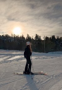 Άτομο που φοράει κράνος και μαύρο εξοπλισμό σκι, κάνει σκι στο χιόνι κοντά σε δέντρα κάτω από συννεφιασμένο ουρανό με τον ήλιο χαμηλά στον ορίζο ντα.