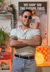 Chemise blanche à manches courtes, jean en denim bleu clair et lunettes de soleil. L'arrière-plan présente une lampe décorative, des plantes sur une étagère et une affiche.