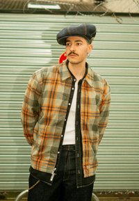 Chemise à carreaux aux teintes gris, orange et bleu, dotée d'une fermeture à boutons et d'un col. Associée à un jean foncé et un béret en denim.