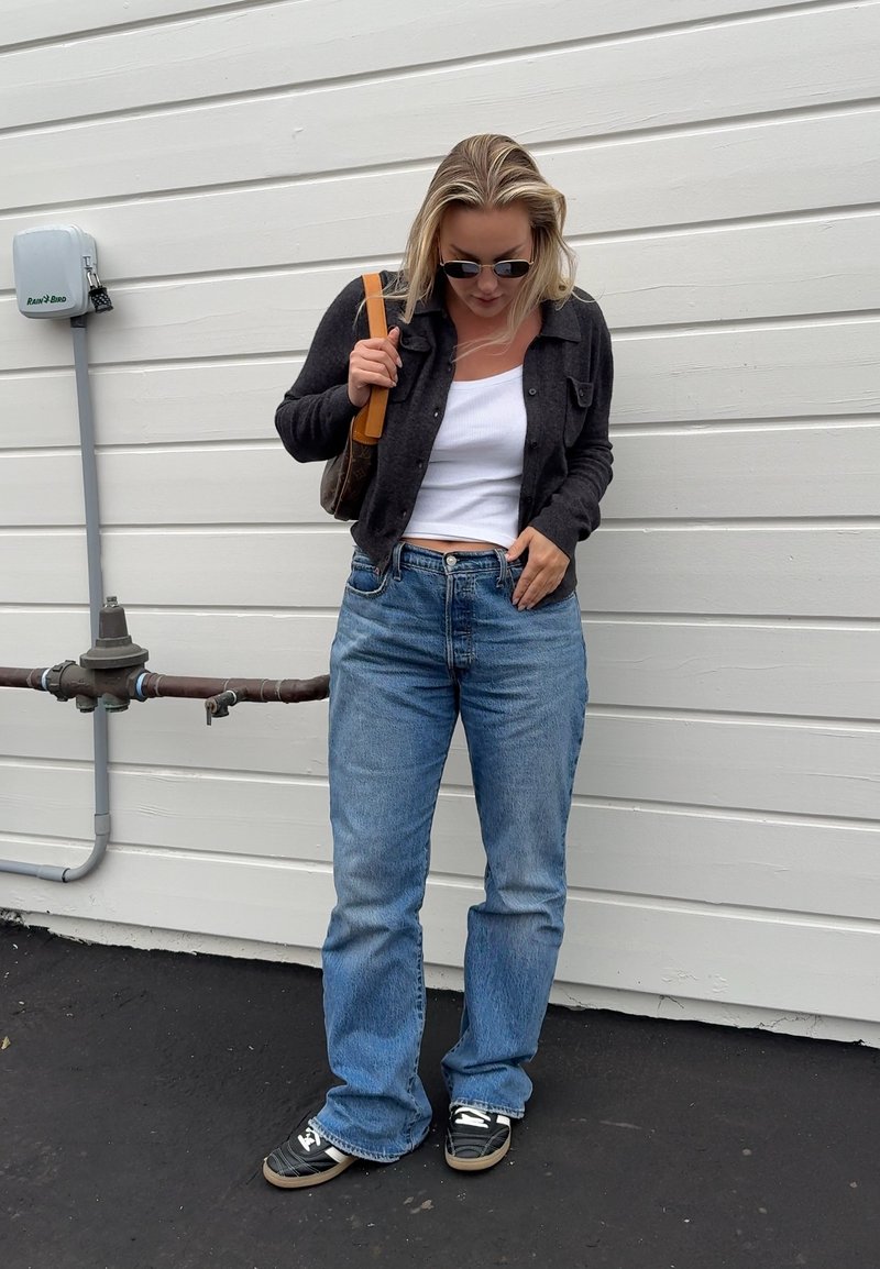 Mujer con gafas de sol, top corto blanco, cárdigan oscuro, vaqueros azules y zapatillas, de pie contra una pared blanca panelada, sosteniendo un bolso marrón de hombro.