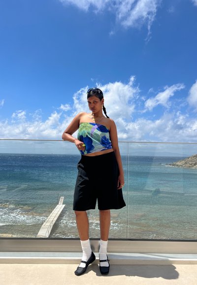 Top corto con estampado floral colorido, pantalones cortos negros, calcetines blancos y zapatos negros con hebillas. Entorno al aire libre junto al mar bajo un cielo azul.
