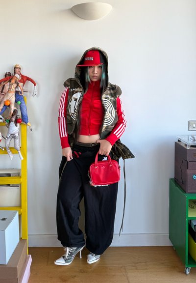 Persona con chaqueta y gorra rojas de Adidas, chaleco con capucha de camuflaje, pantalones anchos negros, zapatillas blancas de caña alta, sosteniendo un pequeño bolso rojo de Adidas en el interior.