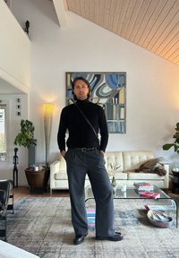 Black turtleneck jumper, grey pinstriped trousers, and chunky black shoes. Indoor setting with a cream sofa, glass coffee table, and artwork.