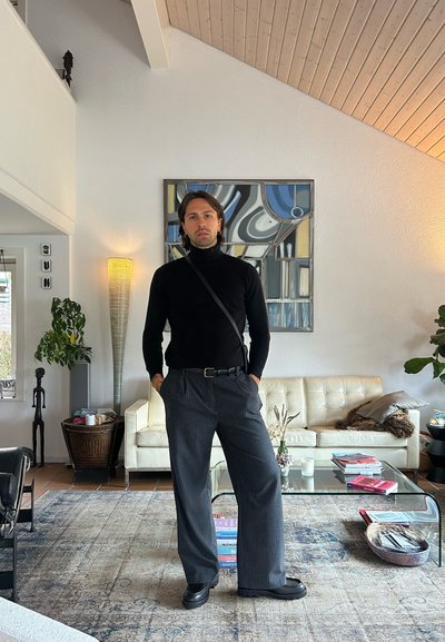 Suéter negro de cuello alto, pantalones de rayas grises y zapatos negros gruesos. Entorno interior con un sofá color crema, mesa de café de vidrio y obras de arte.