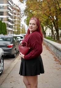 Bordový pletený sveter, čierna rozšírená sukňa, drží malú bordovú kabelku. Pozadie zobrazuje zaparkované autá a jeseňou zafarbené listy.