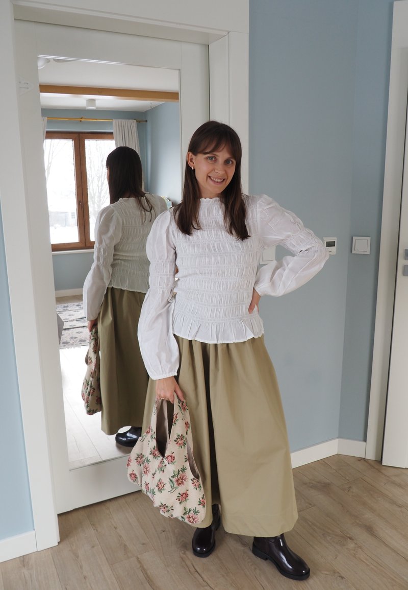 Femme en blouse blanche froncée, jupe beige et bottes noires, souriante, tenant un sac fourre-tout floral près d'un miroir pleine longueur à l'intérieur.