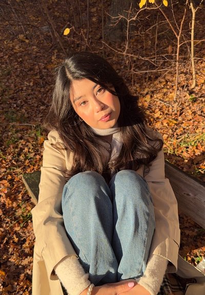 Mujer joven con cabello largo y oscuro sentada en un banco en otoño, abrazando las rodillas, vistiendo un abrigo beige y jeans azules, rodeada de hojas caídas.