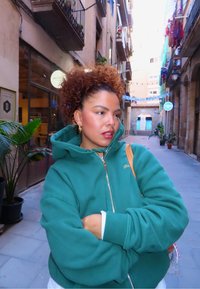 Grønn hettegenser med glidelås og avslappet passform, med ribbestrikkede mansjetter og gullbroderi. I bakgrunnen er en smal gate med planter og bygninger.