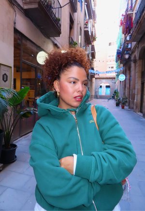 Sudadera con capucha verde con cremallera, ajuste relajado y puños acanalados, con bordado en dorado. De fondo, una calle estrecha con plantas y edificios.