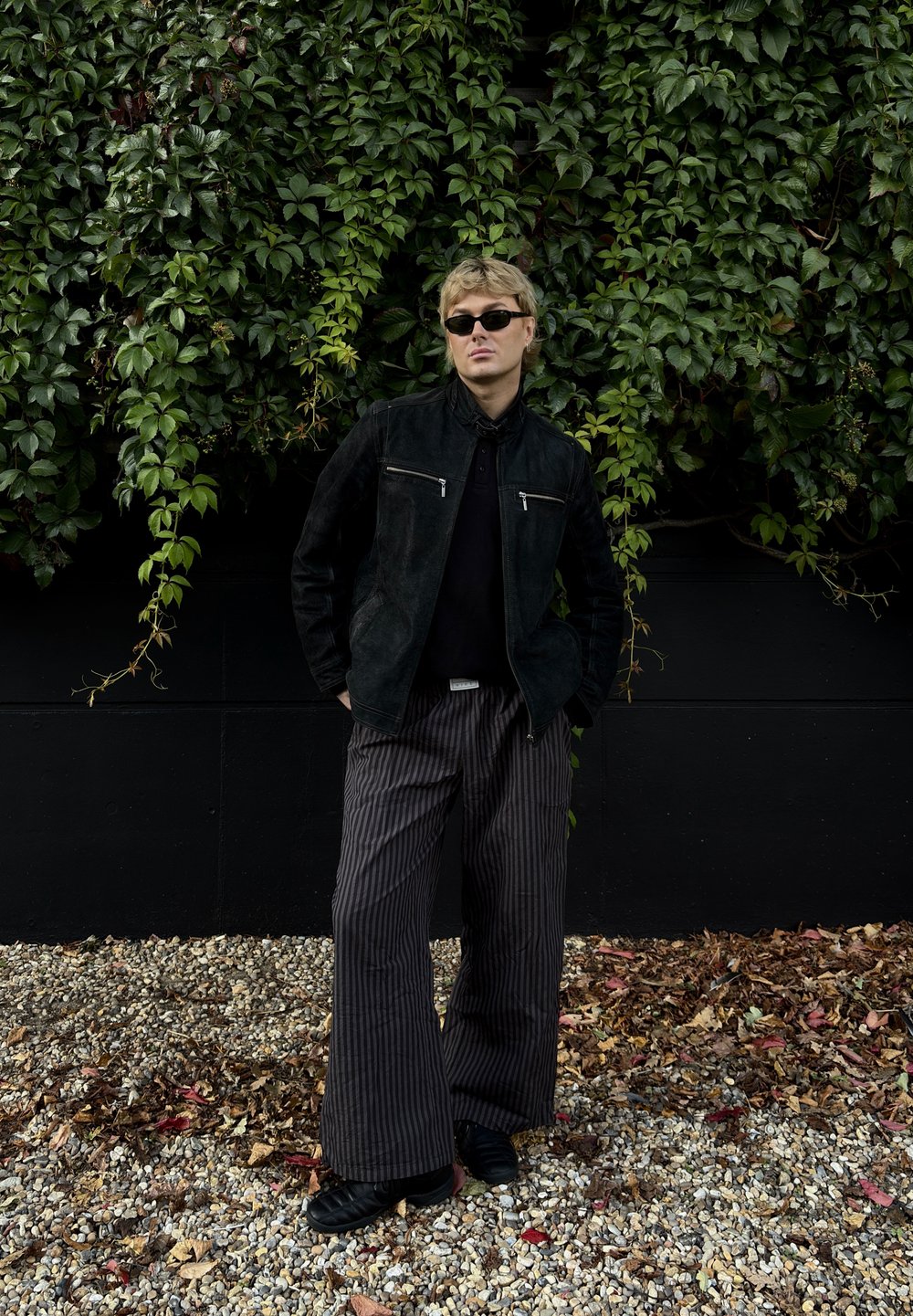 Chaqueta de ante negra con dos bolsillos con cremallera plateados, llevada sobre una blusa negra, combinada con pantalones anchos de rayas blancas y negras y zapatos negros.