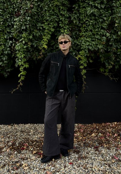 Chaqueta de ante negra con dos bolsillos con cremallera plateados, llevada sobre una blusa negra, combinada con pantalones anchos de rayas blancas y negras y zapatos negros.