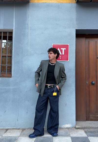 Hombre con cabello rizado viste un blazer gris, camisa negra, jeans anchos oscuros y zapatos azules, de pie contra una pared azul cerca de una puerta de madera y una ventana con rejas.