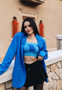 Jeune femme aux longs cheveux foncés portant un haut en tricot bleu court, un blazer bleu vif, une jupe plissée noire et des bijoux en or s'appuie sur une rambarde en pierre à l'extérieur.