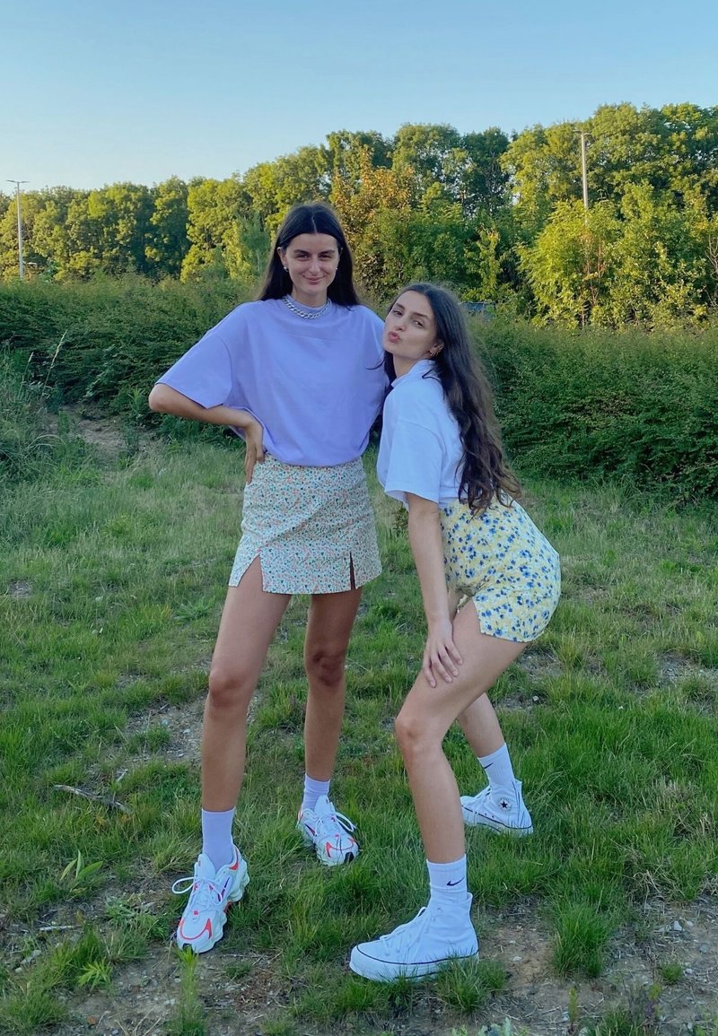 To personer poserer udendørs. Den ene har en lyslilla oversized t-shirt og en blomsteret mini-nederdel på; den anden har en hvid t-shirt og en gul blomsteret mini-nederdel. Begge har hvide sneakers.