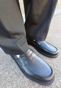 Black leather loafers featuring intricate stitching, a smooth finish, and a slightly elevated sole, paired with pinstriped trousers.