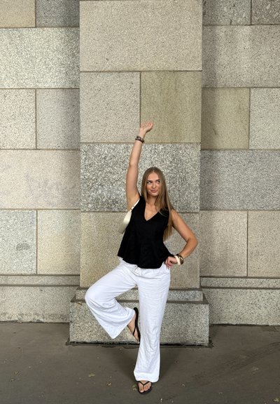 Top negro sin mangas con escote en V, combinado con pantalones anchos blancos. Un bolso de hombro y pulseras acentúan el look. El fondo presenta paredes de piedra texturizadas.