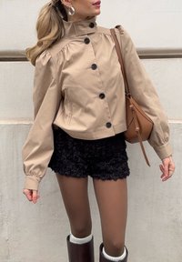 Veste beige courte avec un col haut, boutonnée sur le devant, et manches bouffantes. Short noir à texture florale, leggings couleur tan, et bottes marron.