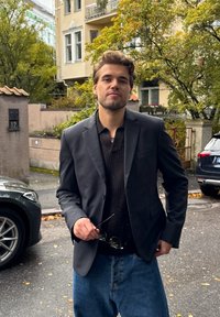 Blazer noir sur un pull en laine sombre, associé à un jean baggy bleu clair. Tenant des lunettes de soleil, debout à l'extérieur avec des arbres en arrière-plan.