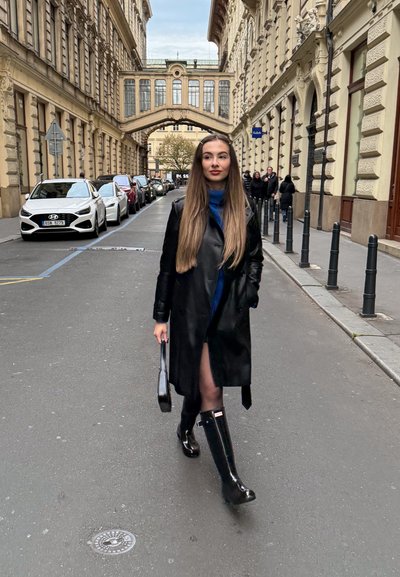 Mujer con cabello largo castaño, vestida con un abrigo de cuero negro y botas, caminando por una calle de la ciudad con edificios históricos y coches aparcados.