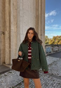 Jonge vrouw met lang bruin haar draagt een groene jas, een rood gestreept topje en een leren rok, terwijl ze een donkerbruine handtas vasthoudt bij een stenen muur.