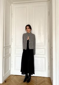 Cardigan gris en tricot avec col montant et fermeture à boutons, superposé à une robe longue noire fluide. Des bottines noires complètent la tenue.