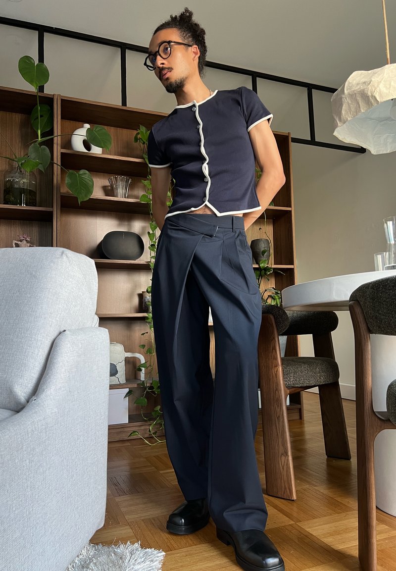 Personne avec des lunettes portant une chemise bleu marine et un pantalon à jambes larges, debout dans un salon avec des étagères en bois et des plantes à l'arrière.