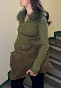 Olivově zelený žebrovaný svetr na zip s chlupatým zeleným límcem; modelka drží hnědou texturovanou pletenou tašku; stojí u schodiště.