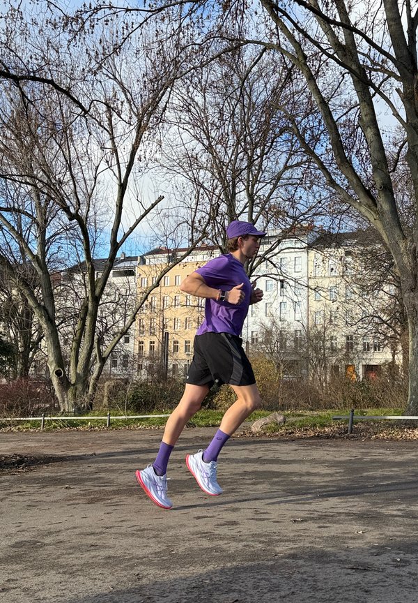 Juoksija violetissa paidassa, hatussa ja sukissa, mustissa shortseissa, valkoiset ja vaaleanpunaiset lenkkarit jalassa, ilmassa polulla, jota ympäröivät puut.
