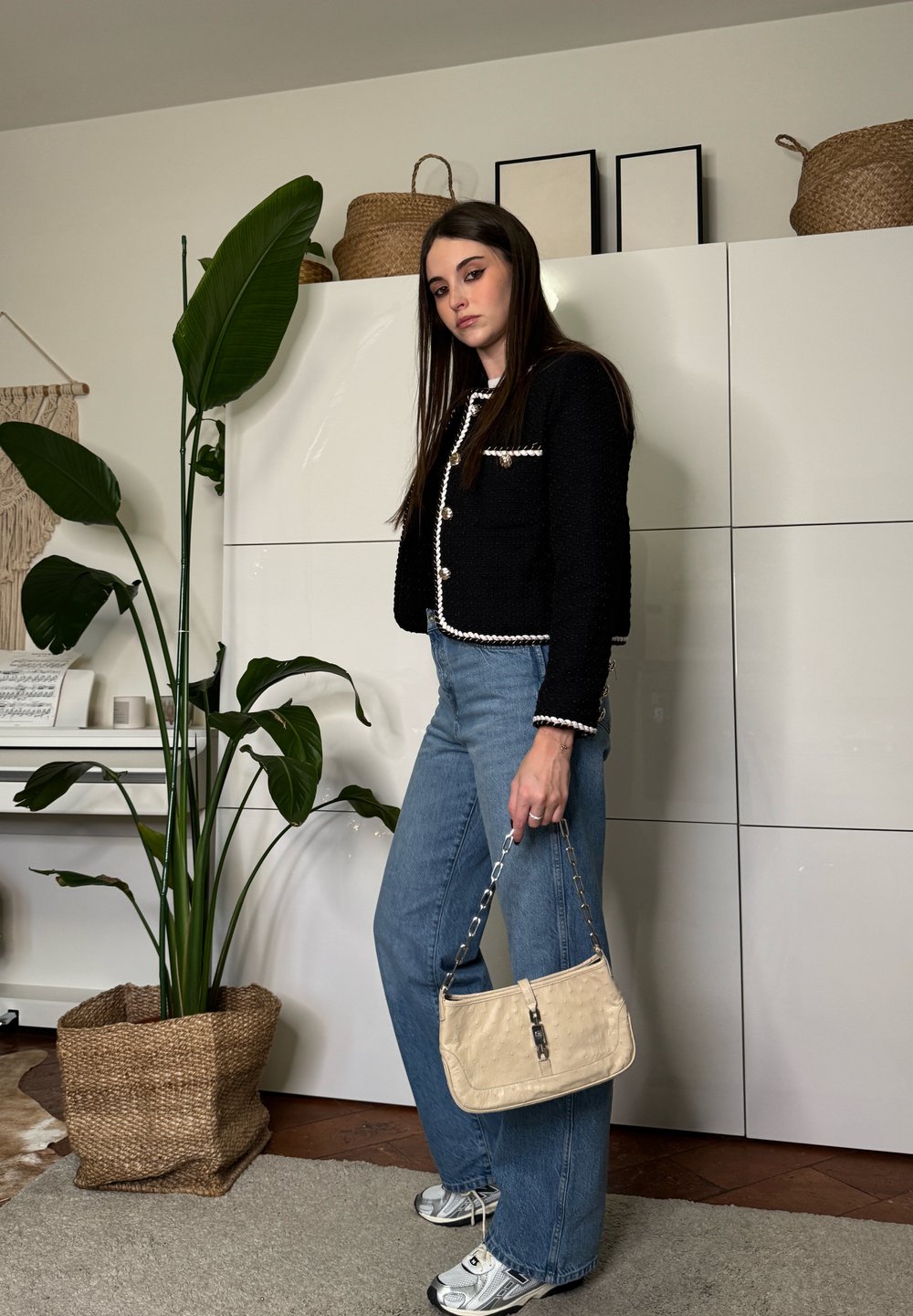 Jeune femme en veste noire et jean tient un sac à main beige, debout à l'intérieur près d'une grande plante verte et de placards blancs.
